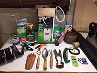 Photo of assorted small garden tools and supplies arranged on table including sprayer, pruning tools, gardening gloves, hose nozzle, and garden liquids.