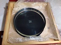 Circular Hubble Telescope lens, black with metallic rim, inside bubble wrap and wooden box.