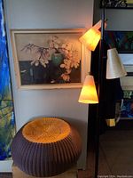 Floor lamp, wicker ottoman, and framed dogwood flower print displayed together, lighting on showing illumination from lamp shades.