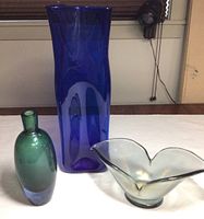 Three glass vases on white surface: tall cobalt blue, green bottle with blue base, and iridescent petal shaped Murano glass vase.