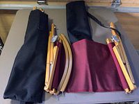 Two unassembled Kermit Chairs laid on table, showing wooden frames, burgundy nylon fabric seats, and black carrying bags with straps.