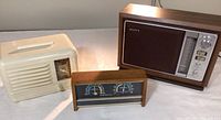 Three items: cream plastic Packard Bell radio, wooden Sony radio, and a wooden barometer side by side.