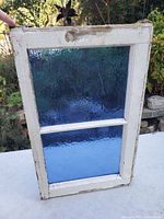 Antique wooden window frame with rippled blue glass panels displayed vertically showing wear on paint and textured blue glass.