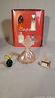 Boxed set of four Calvin Klein mini perfumes in a red box, vintage perfume bottles in front: pink glass with stopper, green Elizabeth Arden bottle with gold bow stopper, and Miss Dior miniature bottle.