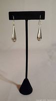 Front view of the pair of elongated water drop shaped silver earrings displayed on a black stand.