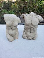Two nude torso sculptures displayed outdoors on a white surface with natural background. Both sculptures have plaster top coating showing cracks and red clay base visible.