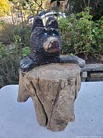 Front and side view of a wooden bear carved from a single piece of wood, partially painted black with blue eyes and red details on the face, sitting on a natural wooden base with cracks and wood texture visible.