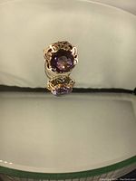 Front view of ring on reflective surface showing faceted synthetic amethyst and openwork gold setting