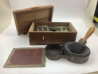 Open view of wooden box showing brass stencil letters inside, metal mortar, wooden pestle, and instruction sheet laid out in front.