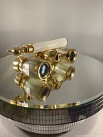 Gold tone opera binoculars with mother of pearl on barrels and extending handle placed on mirrored reflective surface showing front and top details.