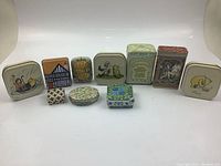 Front view of 10 miniature decorative vintage tins arranged in a single line, showing variation in size, shape, and illustrated designs including animals and flowers.
