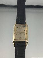 Front view of rectangular yellow gold watch head with cream dial and black leather strap