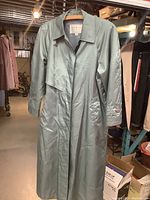 Front view of vintage London Fog trench coat in muted green-grey hanging on hanger showing collar, front button closure, storm flap and cuff strap.