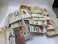 Photo shows a pile of vintage sewing pattern envelopes with illustrations of women and children wearing dresses and skirts. The patterns appear neatly stacked and are mostly in paper packaging.