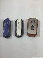 Photo showing three vintage pocket-sized Swiss tools: blue and white Victorinox Swiss Army knives with black sheaths and one silver multi-tool in a brown leather pouch.