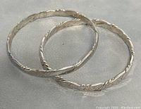 Two round silver bangles with twisted embossed pattern, positioned overlapping in photo on a white leather surface.