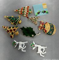 Top view showing the front of the eight Christmas-themed brooches and earrings including Christmas trees, holly leaf, ornament, and white reindeer designs.