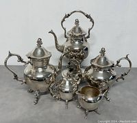 Full view of the entire silver tea set including the teapot with burner and four accompanying pieces.