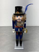Front view of the nutcracker figure showing blue uniform, black top hat with feathers, detailed face with blue eyes, beard, and gold staff on white base.