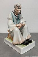 Front-left angle of seated old man figurine holding a whittling knife on a wooden stool, detailed porcelain with painted vest and shoes.