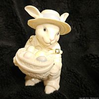 Front view of white porcelain rabbit figurine wearing straw hat holding basket with pastel Easter eggs.