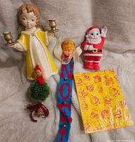 Wide shot showing all five items: large angel figurine holding candlesticks (one detached), small angel with NOEL banner, waving Santa Claus figure, elf doll on green pom pom with bells, and yellow Christmas patterned paper.