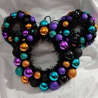 Full frontal image of the black tinsel wreath shaped like Mickey Mouse's head and ears. Decorated with metallic and glitter ornaments in purple, teal, gold, and black.