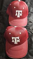 Two maroon Texas A&M hats with mesh fabric and white embroidered 'ATM' logo on front, lying on black background, showing front and top details.