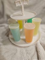 Side view of vintage Tupperware tumblers in pastel colors on white two-tier carousel. One tumbler lid missing.