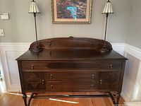 Front view of a dark wooden buffet with three central drawers and two side cabinet doors, standing on curved legs with small caster wheels, placed against a wall with white paneling.