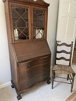 Mahogany secretary desk with upper glass doors, drop-front desk surface, and curved drawers below, next to small ladder back chair with woven seat.