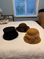 Three wool hats displayed on a white quilted bedspread: black, camel, and chocolate brown.