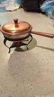 Side angle showing copper lid, brass band around the dish, teak wood handle, and black metal stand with fuel burner underneath.