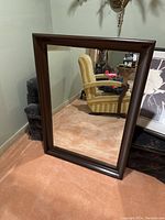 Large rectangular mirror with dark wood frame shown standing against a wall, reflecting a room with a yellow striped armchair and other furniture.