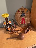 Photo showing assorted native handcrafted art items on wooden surface: carved wooden female figure wearing colorful attire and holding yellow bowl, woven oval wall hanging with a small wooden figure and fabric clothes, small wooden canoe with human figures, carved wooden boat with seated figures, and round rum bottle with rope netting and inscription.