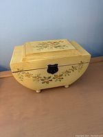 Front and top view of a yellow painted wooden box showing the floral design and curved shape with small feet.