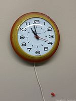 Full view of the retro electric wall clock showing the yellow to orange gradient bezel, white clock face, black numbers, and attached power cord.