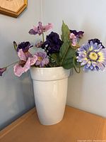 Front view of the tall white porcelain vase with decorative silk flowers in multiple shades of purple and lavender with green leaves.