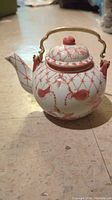 Front angle view of a round white ceramic teapot handpainted with pink floral and vine patterns and brass handle, showing the spout and lid.