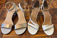 Photo showing three pairs of women's neutral-toned heels on a parquet floor, two pairs white/cream and one pair with metallic silver toe strap.