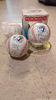 All three Toronto Blue Jays baseballs shown together. One ball is in a Honda-branded clear display case, one ball is loose, and the third is partially wrapped in plastic.