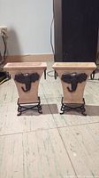 Two beige tapered rectangular planters each featuring a black elephant head relief on the front, placed on black metal stands with curled legs