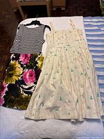 Photo of two summer dresses laid out on a bed, one white with sailboat prints, one striped with floral skirt.