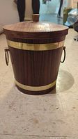 Front view of cone-shaped vintage ice bucket with wood grain and brass bands, featuring a wooden lid knob and brass ring handle.