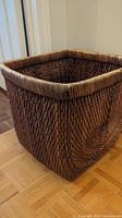 Side and corner view of large square wicker basket placed on parquet floor showing woven pattern and rounded rim.