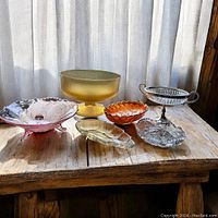 Image showing six vintage glass serving pieces on a wooden table near window light: amber pedestal bowl, pink footed bowl, orange fluted bowl, clear crystal dish, silver and glass pedestal candy dish, and glass leaf plate.