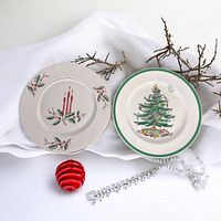 Two Christmas plates together on white fabric with decorative twigs and holiday decor, showing both plate designs.