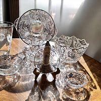 Five glass serving pieces with silver overlay floral designs arranged on table with sunlight, showing intricate metalwork and clear glass.