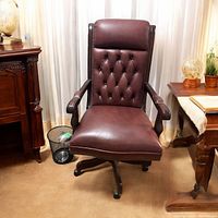 Front view of burgundy leather Chesterfield office chair with mahogany wood arms and base, white gooseneck lamp on side table, and black mesh wastebasket beside it.