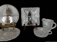 Crystal glass items arranged on black background including a large round bowl, square dish, oval dish, small footed bowl, and handled creamer cup.